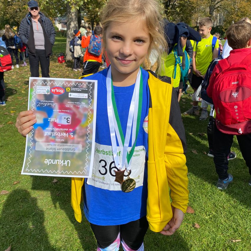 Ein junges Mädchen hält ein Zertifikat und eine Medaille und lächelt für die Kamera. Dahinter sind andere Teilnehmer eines Laufwettbewerbs zu sehen, einige tragen Rucksäcke. Bäume und Gras umgeben das Gebiet.