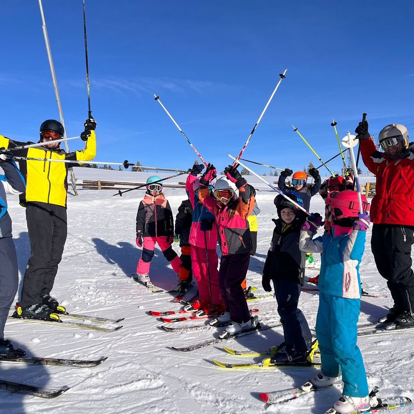 A group of skiers, including adults and children, are on a snowy field. They are holding their ski poles up. Some are wearing goggles and helmets. The snow is white, and the sky is blue.