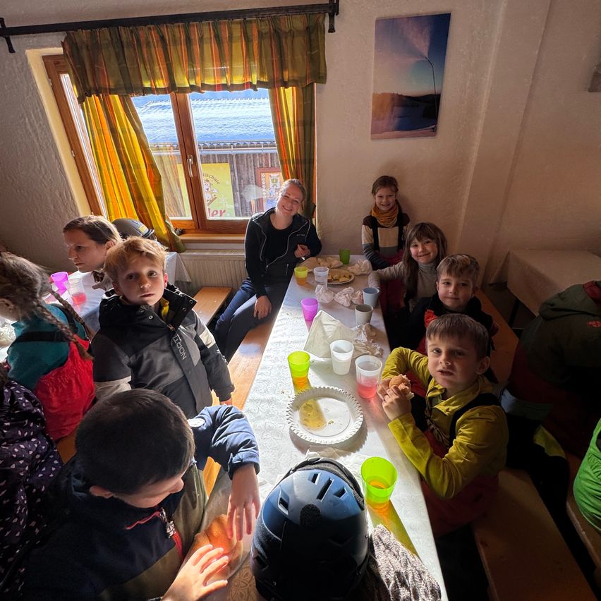 A group of children and adults sitting around a long table inside a room with a window. Some of them are eating while others are holding cups. The table is covered with a white tablecloth and has plates, cups, and a helmet. A woman is sitting on a bench and smiling.