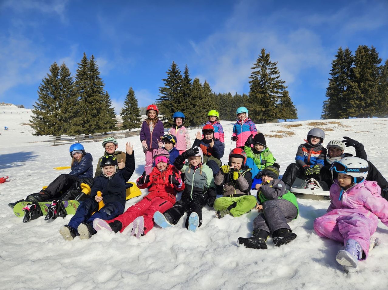 A group of children in winter clothing and helmets sit on the snow with pine trees in the background. They are smiling and posing for a photo.