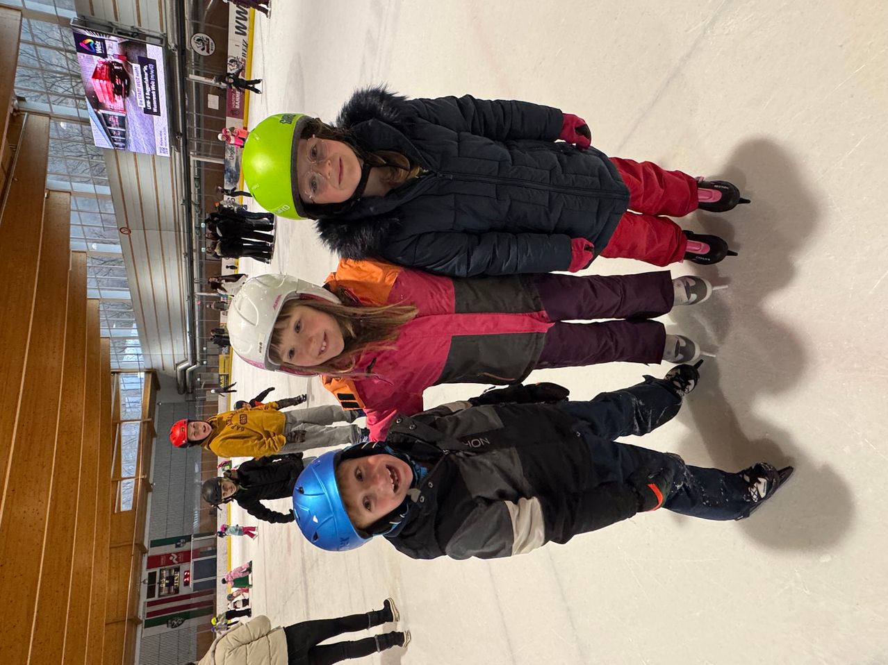 Several children are standing on an ice rink wearing helmets and smiling, with a person standing behind them in the background.
