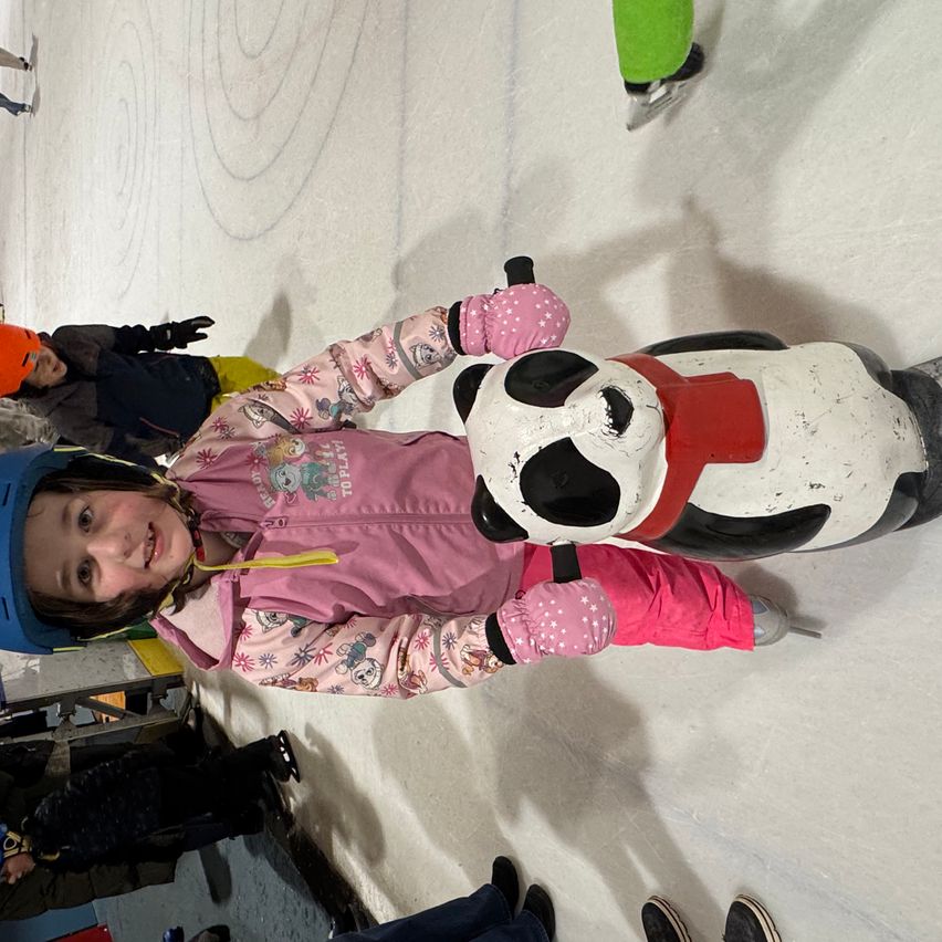 A young girl in pink winter gear holds onto a panda-shaped ice skate aid at an ice rink, with other children and adults visible in the background.