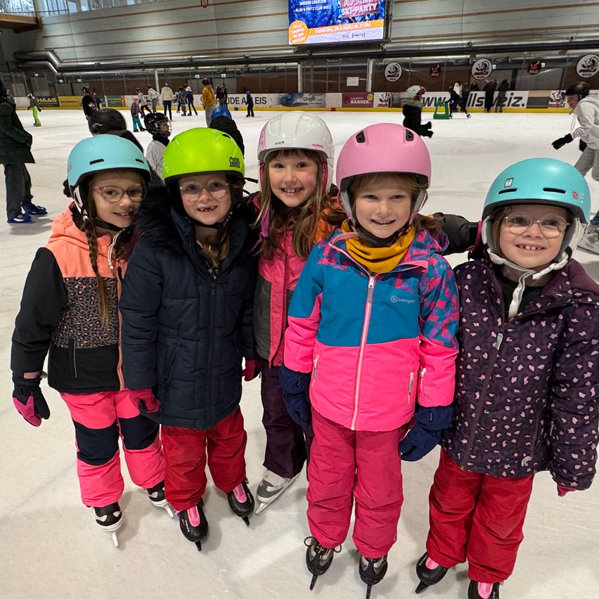Five young girls in winter gear and helmets pose for a photo at an indoor ice rink, with an ice hockey game being advertised on the screen.