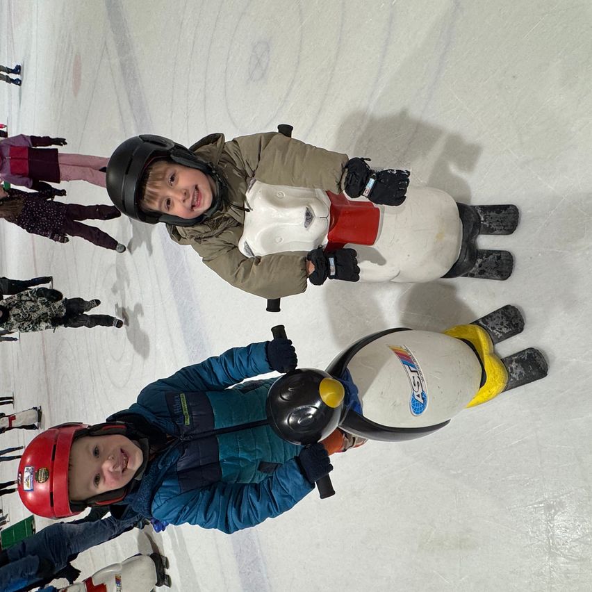 Two young boys ride penguin-shaped scooters on an ice rink, wearing helmets and gloves. Behind them, more children skate and stand, with shadows cast on the ice.