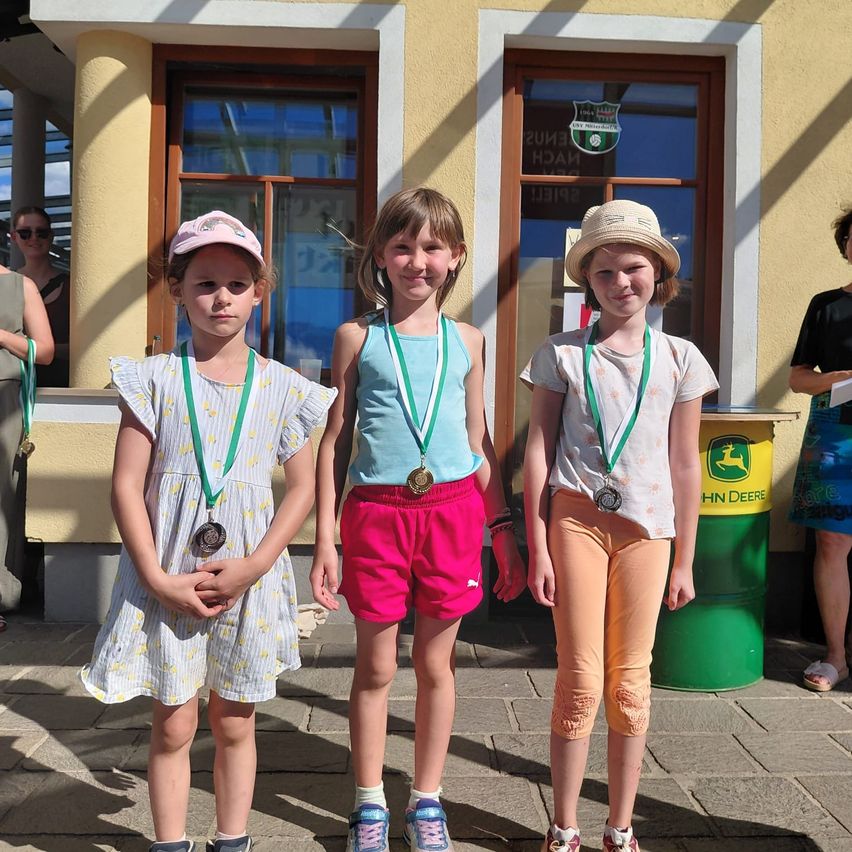 Three girls stand together, each holding a medal, in front of a building with glass doors and a logo. They are wearing caps and sneakers. Behind them are two people, one of whom is holding a piece of paper.