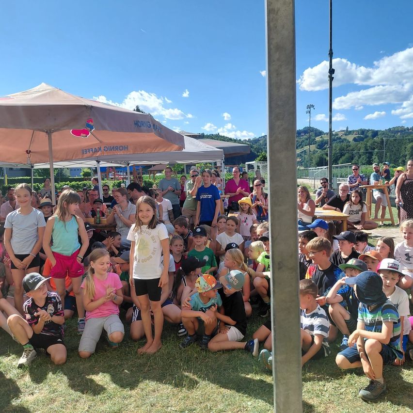 A group of children gathered under umbrellas in a field. Some sit on the grass, while others stand, all facing forward. Behind them are tables, a crowd, and a clear sky with mountains in the distance.