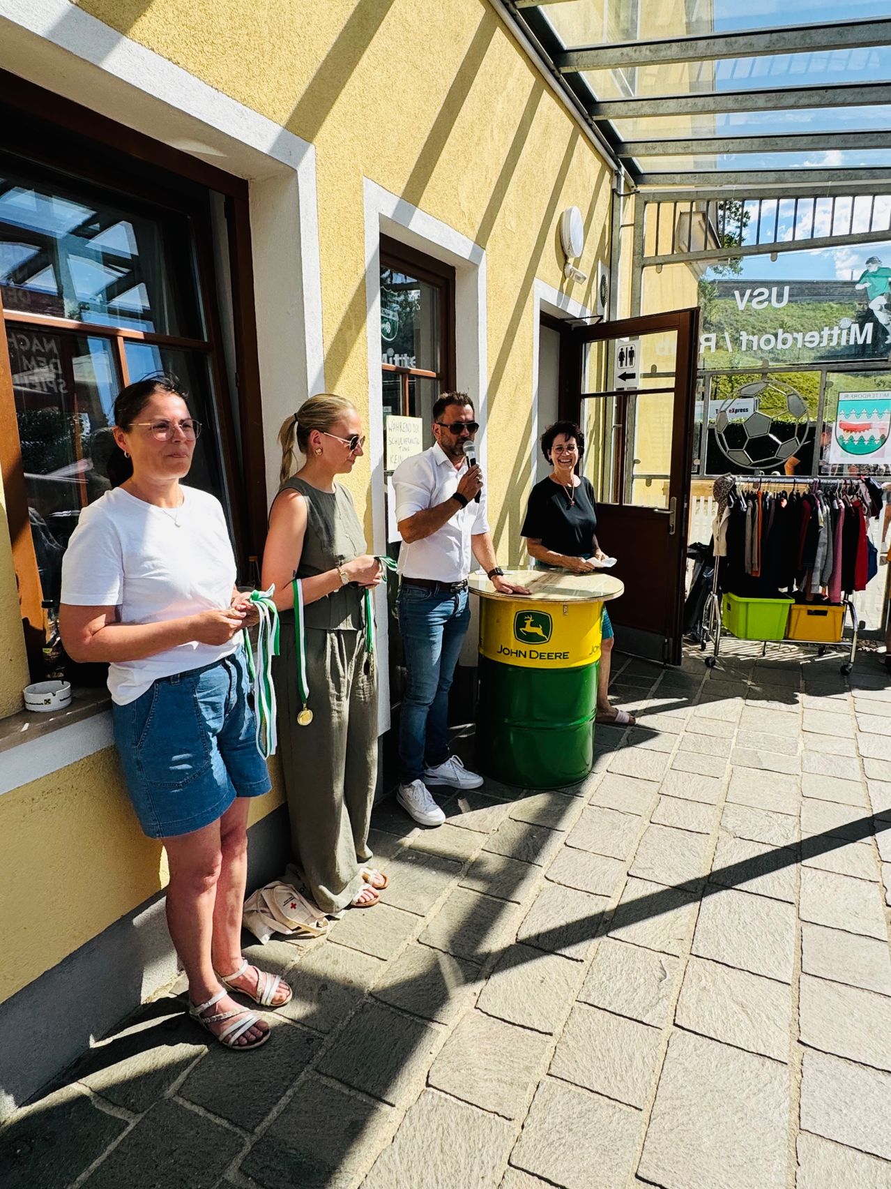 Four people stand in front of a building with a John Deere barrel. A man holds a microphone. The women wear medals. Clothes hang on racks outside the entrance.