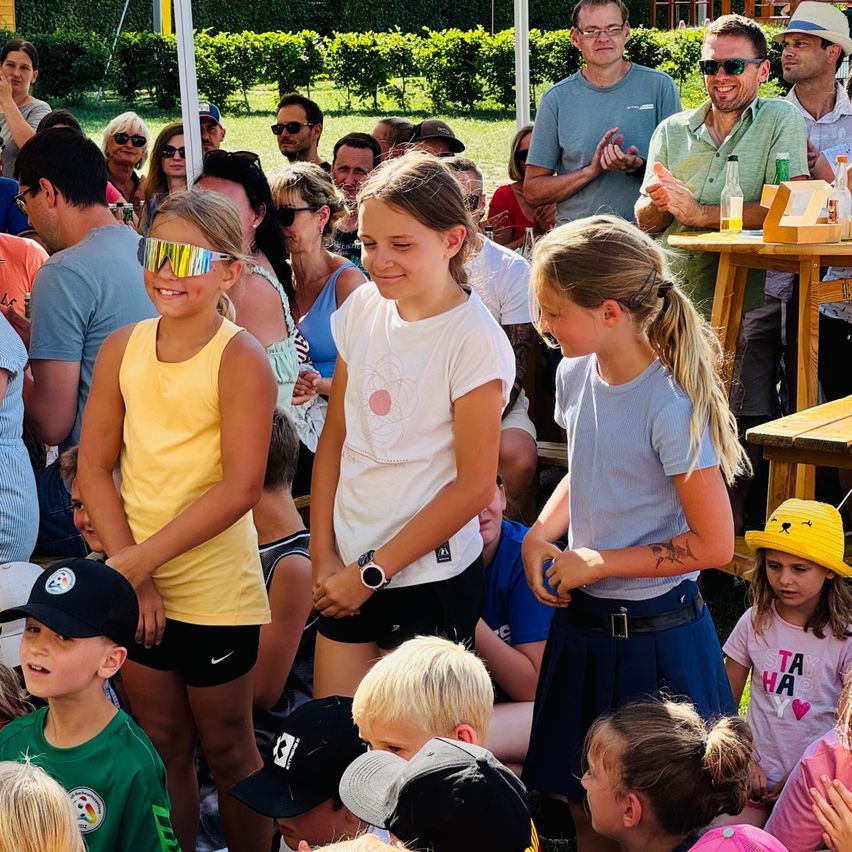 A group of children and adults are standing and sitting outside. Two girls in front wear goggles and dresses. Adults clap in the background.