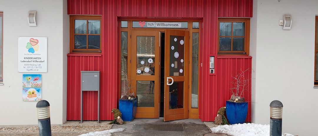 Der Eingang eines Kindergartengebäudes mit roter Außenfassade und einem verschneiten Weg, der zur Tür führt. Auf beiden Seiten des Eingangs stehen zwei Topfpflanzen.