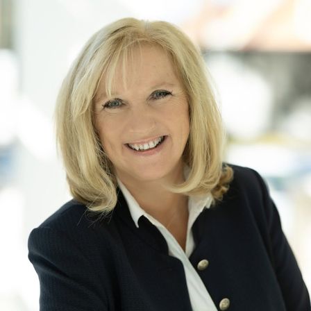 Eine Frau mit kurzen blonden Haaren lächelt und schaut nach vorne. Sie trägt ein weißes Hemd und eine schwarze Jacke mit goldenen Knöpfen. Der Hintergrund ist unscharf.