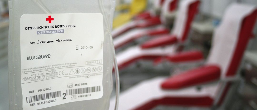 Ein Nahaufnahme einer Blutspendenbeutels mit einem Roten Kreuz Symbol. Er enthält 287 Milliliter Blut. Dahinter sind mehrere rote Stühle zu sehen, wahrscheinlich in einer Blutspendestation.