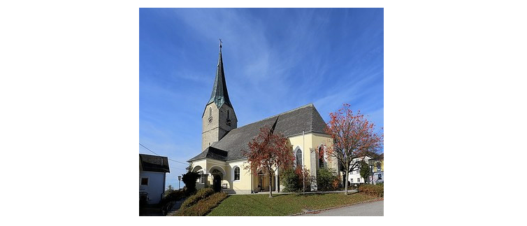 Eine kleine Kirche mit einem Turm und einem grauen Dach steht auf einem Hügel unter einem blauen Himmel.