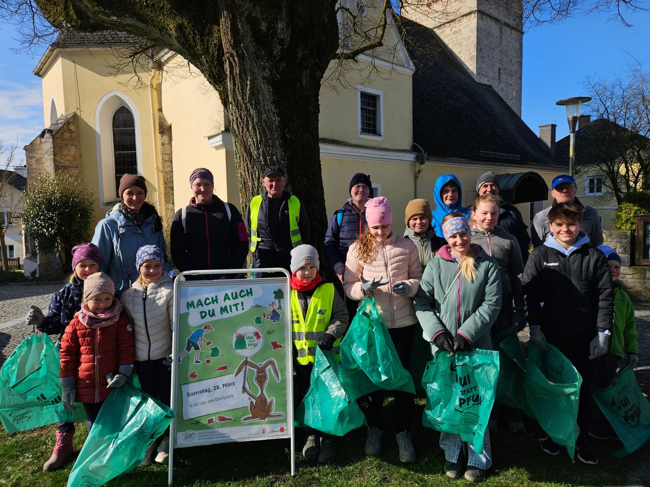 Eine Gruppe von Menschen, einige in Warnwesten, steht vor einer Kirche und hält grüne Beutel. Sie posieren für ein Foto. Der Banner liest 'MACH AUCH DU MIT!'. Das Datum ist der 28. März.