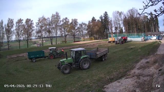 Mehrere Traktoren und Anhänger sind auf einem Rasenfeld geparkt. Das Datum und die Uhrzeit lauten 09/04/2026 08:29:13. Im Hintergrund befinden sich Bäume und ein Zaun.