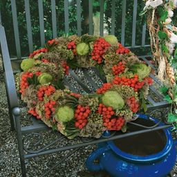Ein Kranz aus roten Beeren und grünen Blättern ist auf einer Metallbank ausgestellt, darunter ein blauer Topf.