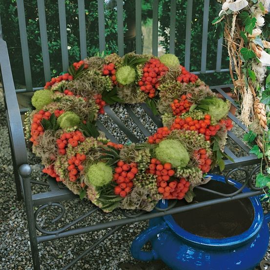 Ein Kranz aus roten Beeren und grünen Blättern ist auf einer Metallbank ausgestellt, darunter ein blauer Topf.