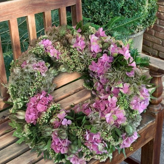 Ein Blumenkranz mit rosa Hortensien und grünen Blättern auf einer Holzbank, mit einer Ziegelmauer und einem Topfpflanzen im Hintergrund.
