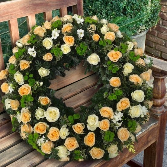Ein Kranz aus gelben und weißen Rosen mit grünen Blättern liegt auf einer Holzbank, mit einem Topfpflanzen im Hintergrund.