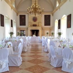 Ein großer Saal mit weiß bezogenen Tischen und Stühlen, geschmückt mit weißen Schleifen und Blumenarrangements. Eine Kristallleuchter hängt von der Decke, und Gemälde hängen an den Wänden.