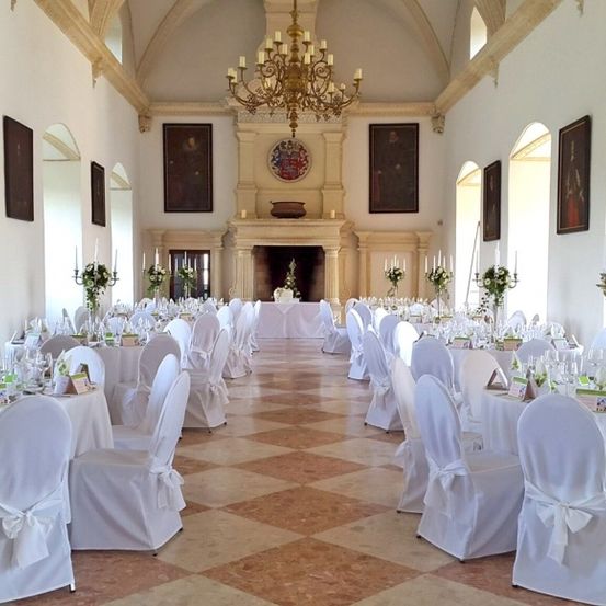 Ein großer Saal mit weiß bezogenen Tischen und Stühlen, geschmückt mit weißen Schleifen und Blumenarrangements. Eine Kristallleuchter hängt von der Decke, und Gemälde hängen an den Wänden.