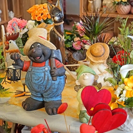 Ein Tisch mit dekorativen Figuren, darunter eine lächelnde Figur mit Axt, umgeben von verschiedenen Blumen, einer Katze und einer Laterne.