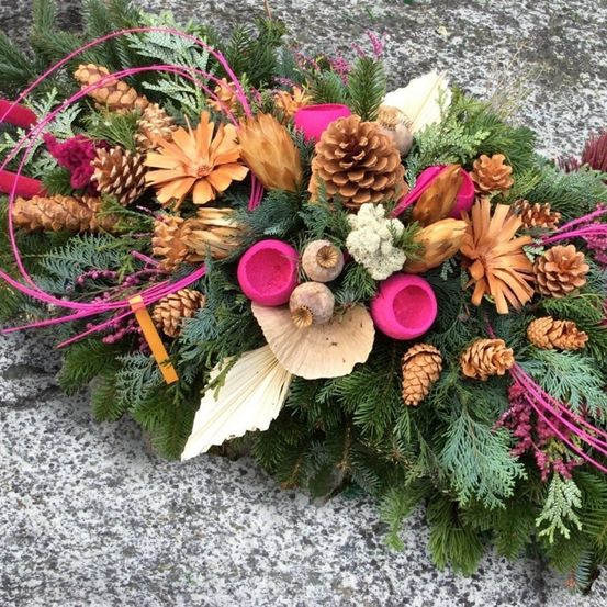 Ein dekorativer Kranz mit einer Mischung aus getrockneten Blumen, Tannenzapfen und Blättern, verziert mit rosa Bändern und roten Akzenten, ruht auf einer Steinoberfläche.