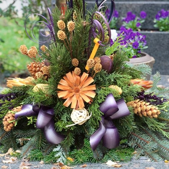 Eine farbenfrohe Blumengestecke mit einer orangefarbenen Daisy, lila Bändern und Tannenzapfen, vor dem Hintergrund von lila Blumen und üppiger Grünpflanze.