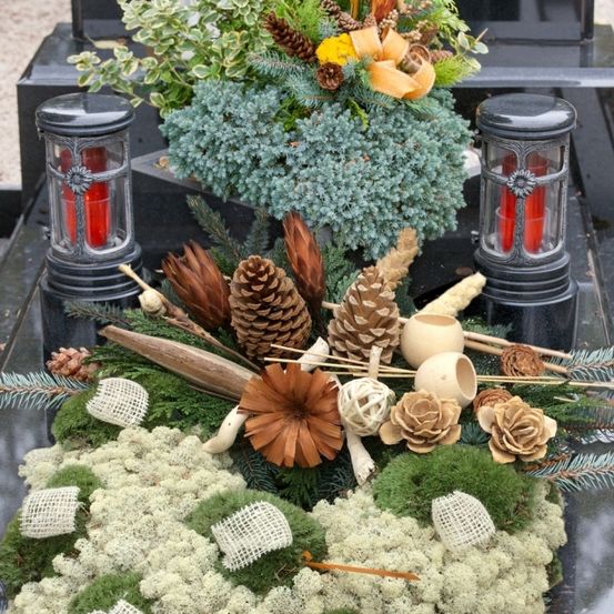Ein Gedenkarrangement mit immergrünen Pflanzen, Tannenzapfen, Flechten und einer roten Kerze auf einer Granitfläche mit zwei Laternen.