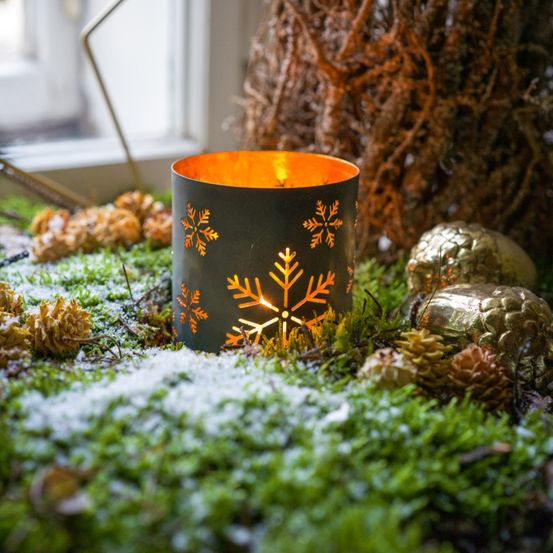 Eine Kerze in einem Metallhalter mit Schneeflocken-Ausschnitten brennt hell auf einem Bett aus Moos, Tannenzapfen und Schnee, mit einem Fenster im Hintergrund.