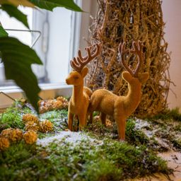 Zwei dekorative goldene Rehe stehen auf einer grünen moosigen Oberfläche mit Tannenzapfen, vor dem Hintergrund eines Strohbaums und eines Blattes.