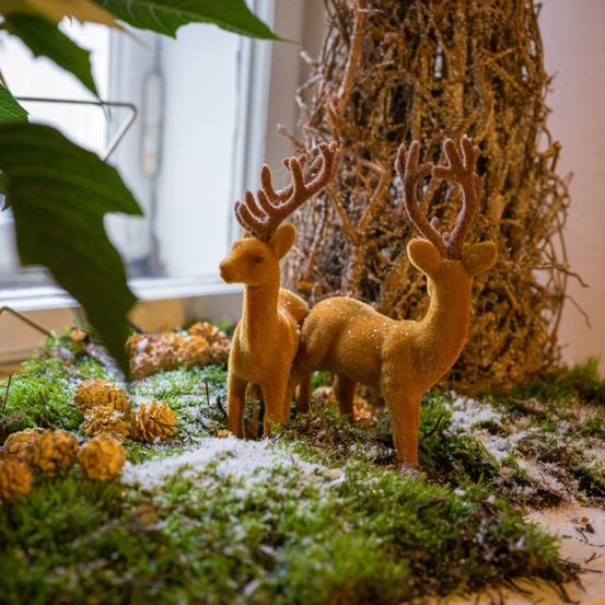 Zwei dekorative goldene Rehe stehen auf einer grünen moosigen Oberfläche mit Tannenzapfen, vor dem Hintergrund eines Strohbaums und eines Blattes.