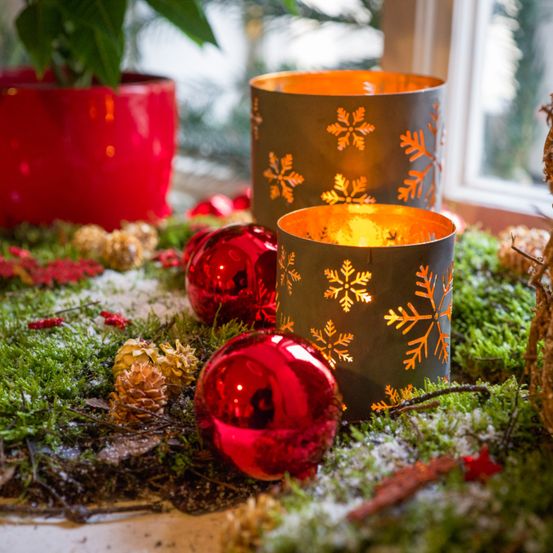 Zwei Kerzenhalter mit Schneeflockenmustern stehen auf einem Tisch, umgeben von Weihnachtsdekorationen, roten Kugeln und Tannenzapfen. Im Hintergrund ist eine rote Topfpflanze zu sehen.