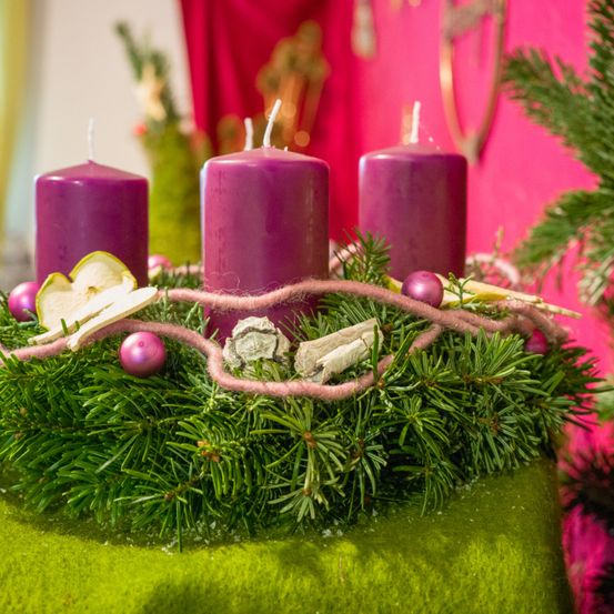Ein Kranz mit drei lila Kerzen und einer rosa Schleife, zusammen mit einem Apfelstück und Weihnachtsdekorationen, steht auf einer grünen Oberfläche.