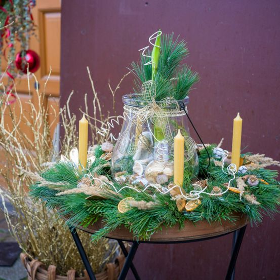 Ein festlicher Kranz mit grünen Blättern, goldenen Strohhalmen und drei gelben Kerzen steht auf einem Holztisch. Er enthält eine Glasvase mit einem Band und einer getrockneten Pflanze darin.