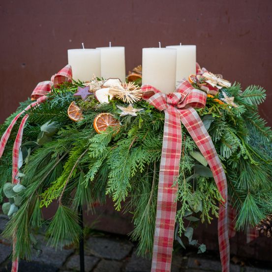 Ein Weihnachtskranz mit vier weißen Kerzen, roten und weißen Bändern und dekorativen Ornamenten vor einem braunen Hintergrund.