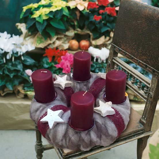 Ein Holzstuhl trägt einen Kranz mit fünf lila Kerzen und zwei weißen Sternen. Im Hintergrund stehen verschiedene Blumen, darunter rote, rosa und gelbe.