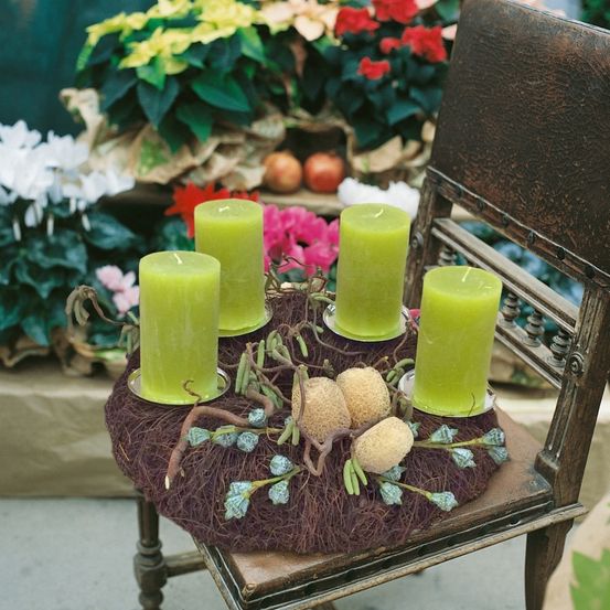 Ein Holzstuhl trägt einen Kranz mit grünen Kerzen und Tannenzapfen. Der Kranz steht auf einer Holzunterlage und ist von Blumen umgeben.