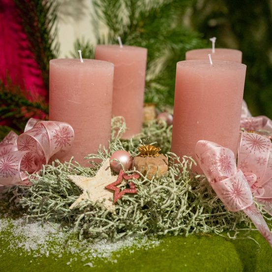 Vier rosa Kerzen, umgeben von dekorativen Gegenständen wie Bändern und Ornamenten, sind auf einer grünen Oberfläche ausgestellt.