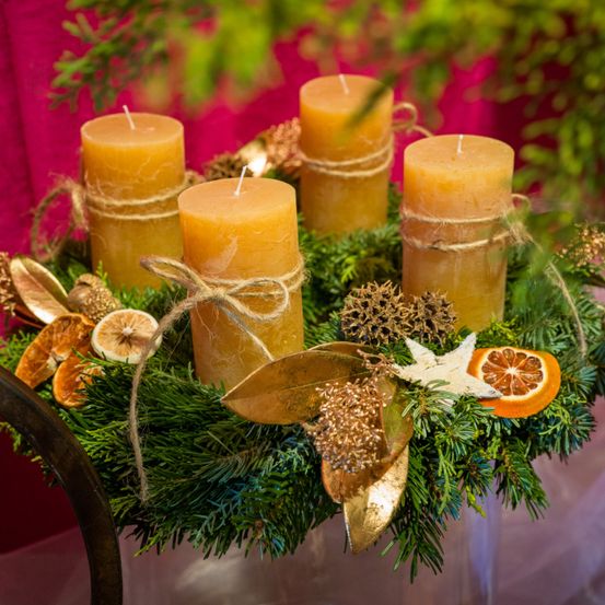 Ein Kranz mit fünf gelben Kerzen, in Twine gewickelt, umgeben von getrockneten Blättern, Seesternen und Orangenscheiben vor einem roten Hintergrund.