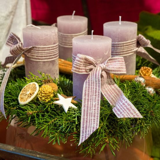 Vier lavendelfarbene Kerzen, mit Bändern gebunden, sind in einem Kranz mit Tannennadeln, Stern und getrockneten Früchten angeordnet. Die Kerzen stehen auf einem Holzstab mit Tannenzapfen.
