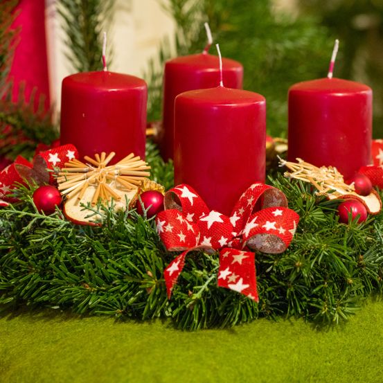 Ein Kranz mit fünf roten Kerzen, geschmückt mit goldenen Ornamenten, roten Sternen und einem roten Band, liegt auf einer grünen Oberfläche. Die Kerzen sind nicht angezündet.