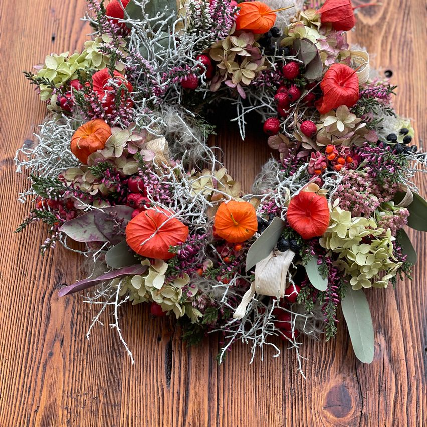 Ein Kranz aus getrockneten Blumen und Blättern, einschließlich roter und orangefarbener Blütenblätter, auf einer Holzoberfläche angeordnet.
