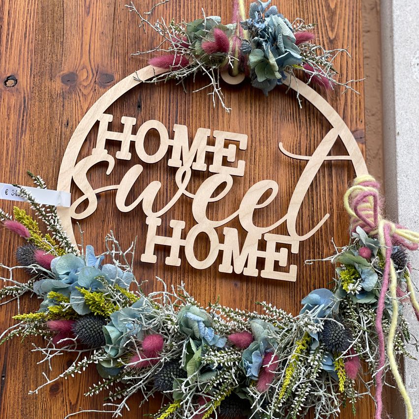Ein hölzerne Girlande mit dem Text 'HOME Sweet HOME' hängt an einer Holztür, geschmückt mit verschiedenen bunten Blumen und Zweigen.