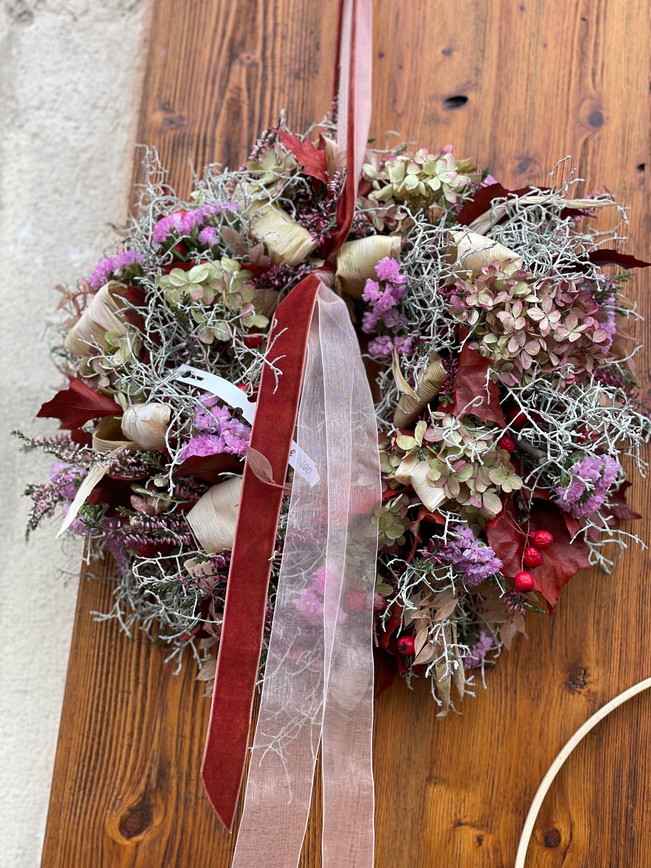 Ein Kranz aus getrockneten Blumen, einschließlich rosa, lila und weißen Blüten, ruht auf einer Holzoberfläche mit einem roten Band.