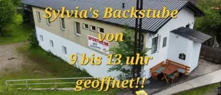 An aerial view of Sylvia's Backstuben, a white building with a black roof, featuring a sign that reads '9 bis 13 Uhr geoffnet' and a bench and table outside.