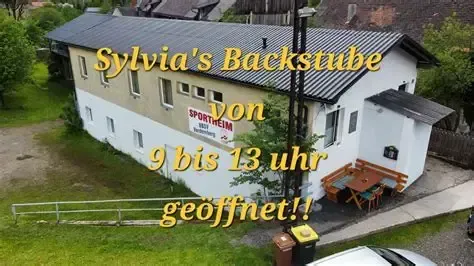 Ein Luftbild von Sylvias Backstuben, einem weißen Gebäude mit schwarzem Dach, das ein Schild mit der Aufschrift '9 bis 13 Uhr geöffnet' und eine Bank und einen Tisch draußen zeigt.