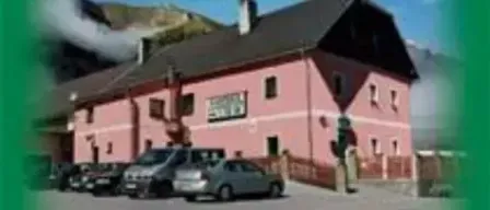 Ein rosa Gebäude mit schwarzem Dach ist im Hintergrund. Mehrere Autos sind davor geparkt. Das Gebäude hat einen Schornstein auf dem Dach und mehrere Fenster.