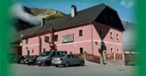 Ein rosa Gebäude mit schwarzem Dach ist im Hintergrund. Mehrere Autos sind davor geparkt. Das Gebäude hat einen Schornstein auf dem Dach und mehrere Fenster.
