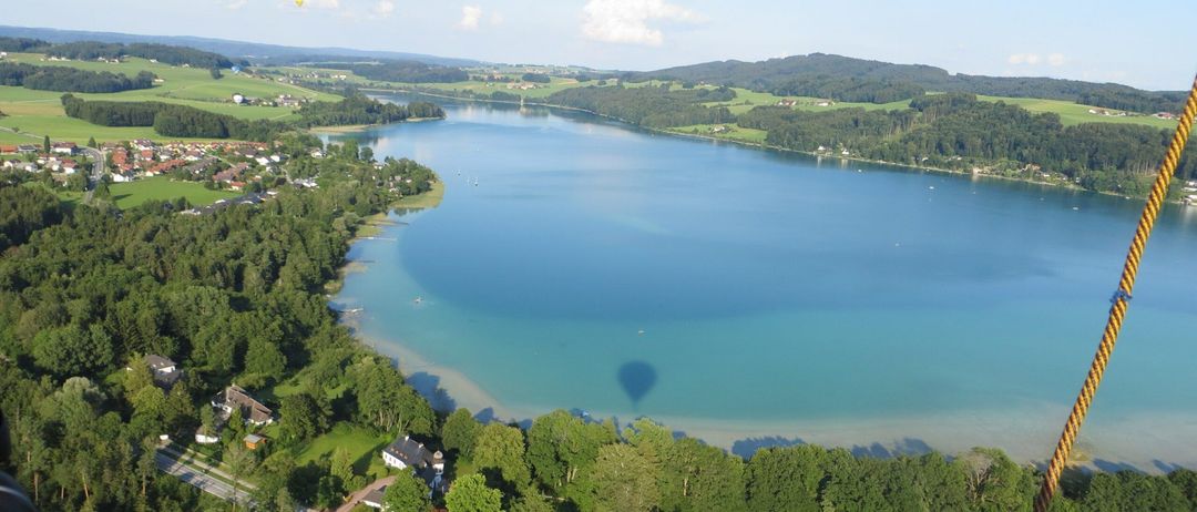 Luftaufnahme eines friedlichen Sees, umgeben von üppigem Grün und entfernten Bergen. Ein Heißluftballon schwebt oben, mit einem kleinen Haus am Ufer.