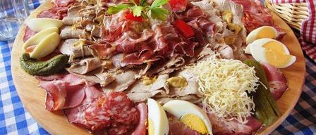 Eine Holzplatte mit Schinken, Eiern, Käse und Gurken auf einer blau-weiß karierten Tischdecke.